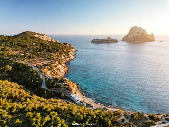Aerial view of a coastal landscape in Ibiza, Balearic Islands, with a winding road, forested cliffs, and rocky islands in the sea at sunset.
