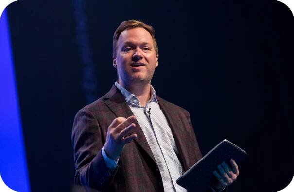 A photo of Jeremy Miller speaking on a stage with a tablet in his hand.