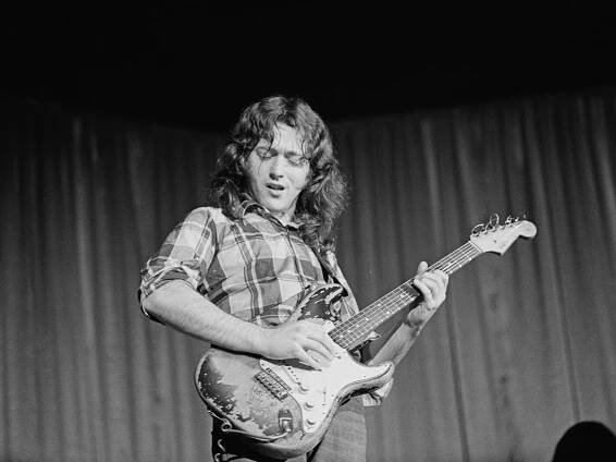 Rory Gallagher pictured in black and white playing his Stratocaster on stage.