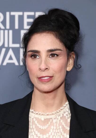 Sarah Silverman in formal attire with intricate lace top and blazer, posing against a backdrop with partial text visible