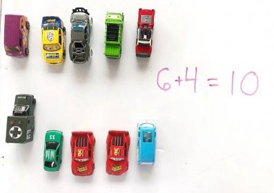Matchbox cars lined up in a group of six and a group of four with the equation 6+4=10 written on a whiteboard in pink.