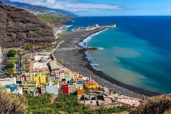 Aerial view of Tazacorte, La Palma, showing a black sand beach, harbor, and colorful buildings.