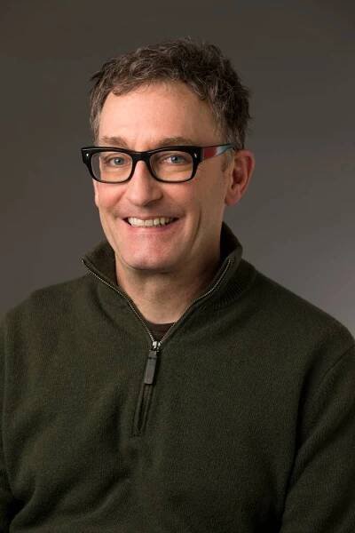 Tom Kenny smiling, wearing glasses and a zip-up sweater, against a neutral background