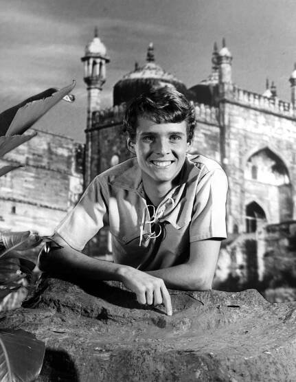 Jay North leaning against a rock with ornate buildings in the background in a scene from the film 'Maya', 1966