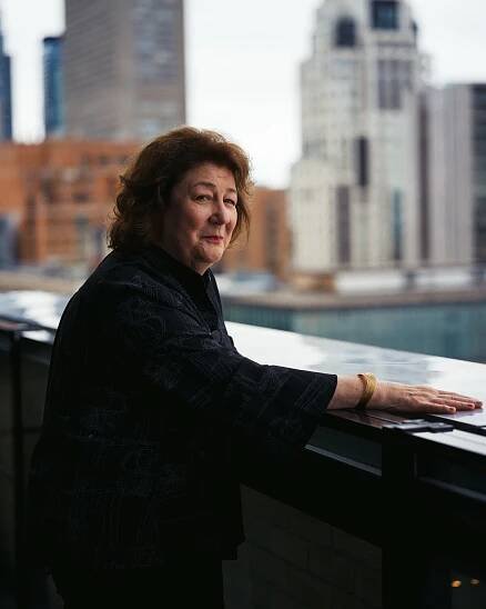 Margo Martindale in a dark blouse stands against the side of a balcony and looks over her shoulder at the camera