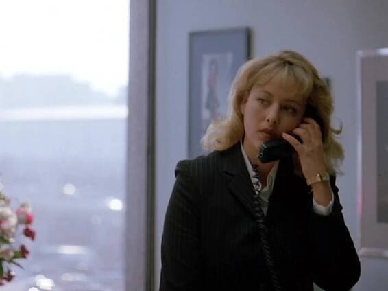 Virginia Madsen as Brenda Hopkins in Full Disclosure, wearing a black pinstripe suit and gold watch. She holds a phone to her ear with a serious expression in a bright office with large windows.