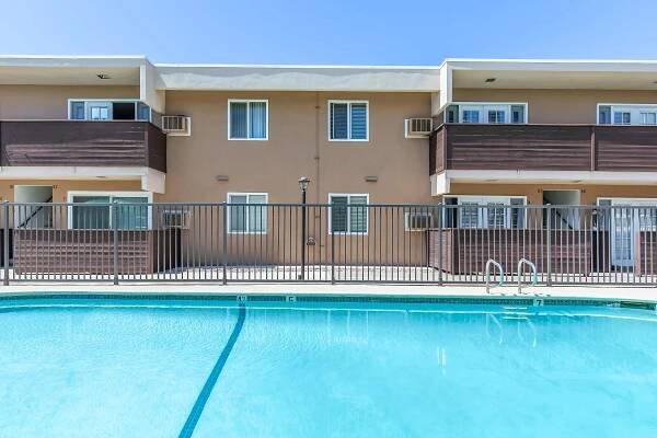 View of an apartment building with balconies, situated next to a swimming pool. The pool is in the foreground, and the building is in the background under a clear blue sky.