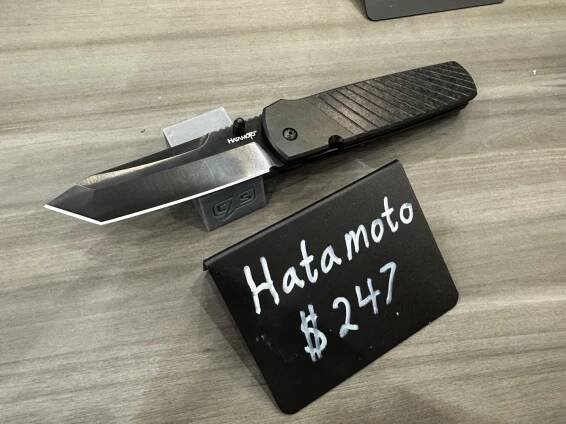 This image shows the Cold Steel Hatamoto folding knife on display at the Cold Steel booth during the 2025 Blade Show. The knife is laid out with the blade open, revealing a 4-inch tanto blade made from CPM 20CV steel, which has a polished finish. The handle is crafted from full carbon fiber, giving it a lightweight yet high-end look. The knife rests on a branded display surface, and there may be a product card or label nearby with specifications. Other Cold Steel products and booth signage are visible in the background, but the Hatamoto is the clear focus of the shot. Lighting from the convention floor highlights the blade’s shape and the texture of the carbon fiber handle.