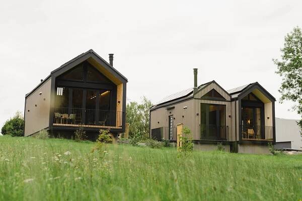 Zwei moderne Tiny Houses aus Holz mit großen Fenstern und Veranden, umgeben von grüner Natur unter einem bewölkten Himmel, individuell konfiguriert als WOLF Häuser.