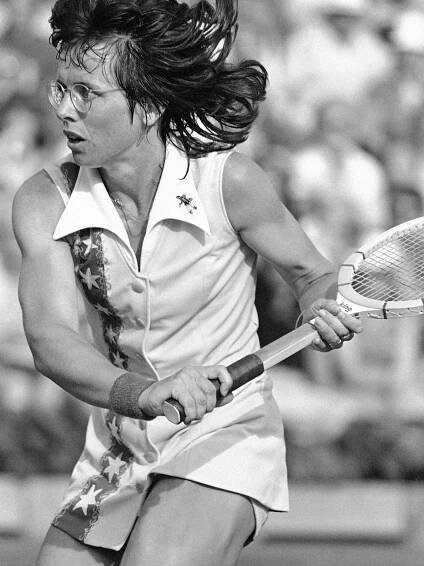 A black and white, 1977 active shot photograph of a muscular young female athlete with glasses, and a white tennis outfit with stars down the right side, preparing for a backstroke with the tennis racket.