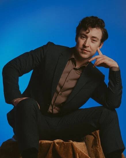 A man in a dark suit and brown shirt sits with one hand on his leg and the other against his face.