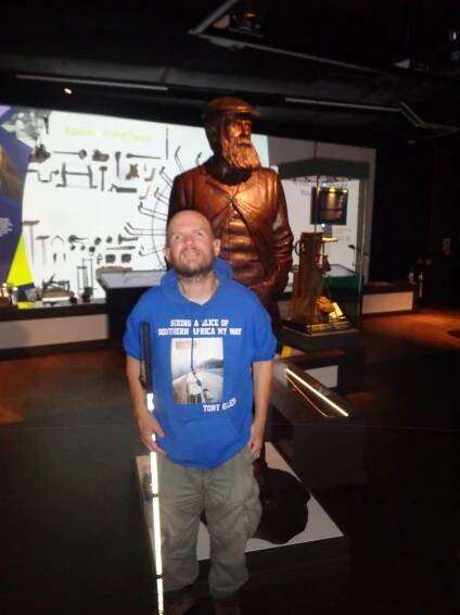Tony in the R&A World Golf Museum. He is at the entrance to the exhibitions with a larger than life bronze statue of Old Tom Morris, the Grand Old Man of Golf, in the background. Thomas Morris (1821 – 1908) was a Scottish professional golfer, greenskeeper  and course designer, born in St Andrews. He is a famous golfing figure noted for winning the Open Championship four times and for designing or remodelling more than 70 golf courses.