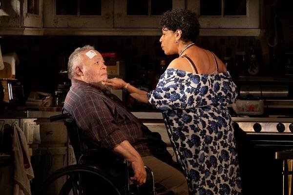 In a scene in “Between Riverside and Crazy,” Stephen McKinley Henderson and Liza Colón-Zayas are facing each other in a kitchen. Henderson is in a wheelchair and has a bandage on his forehead. Colón-Zayas is standing in front of him in a white and blue floral dress, reaching out toward his face.