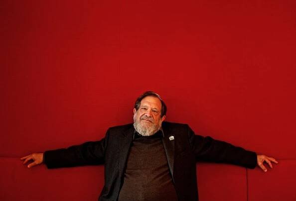 Michael Lerner, a bearded man wearing a yarmulke, leans back on a red sofa against a red wall, his arms outstretched.