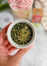 A woman's hand holds a small white ramekin filled with Furikake, showing the individual pieces of dried nori, sesame seeds, and grains of salt and sugar