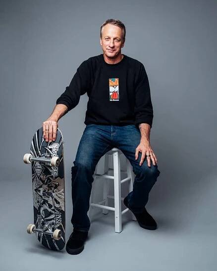 A person sits on a stool wearing casual jeans and a graphic sweatshirt, holding a skateboard upright