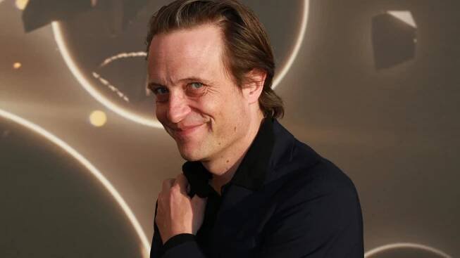 August Diehl steht vor einer Wand auf die Kreis projiziert sind und lacht in die Kamera