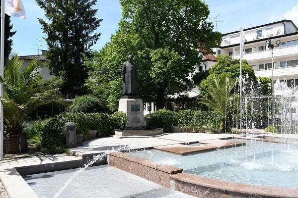 Denkmal mit Springbrunnen und Palmen im Kurpark bei Sonnenschein.