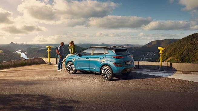 Mladá dvojice sedí na kapotě SUV Hyundai Kona Electric zaparkovaného na místě s výhledem do malebné krajiny.