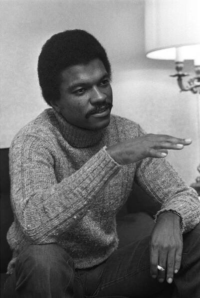 In this black-and-white picture, Billy Dee Williams is wearing a turtleneck sweater and jeans. He appears to be gesturing, midsentence, and he holds a cigarette between the index and middle fingers of his left hand. 