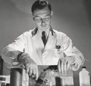 A man in a lab coat uses tongs to take a substance out of a test tube held behind a shield.