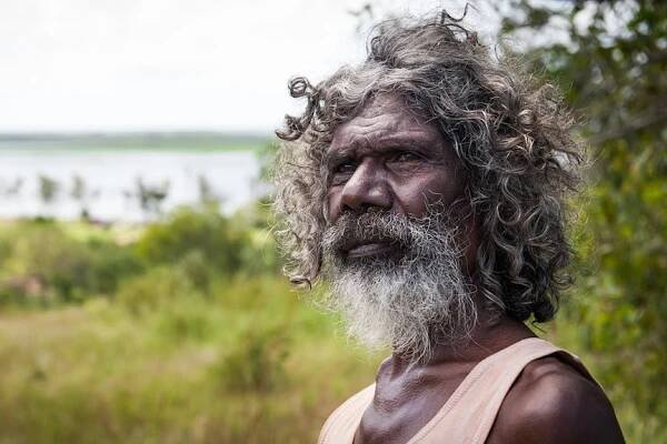 An image of an older Indigenous man, with long hair and a beard, with greenery and a river behind him.
