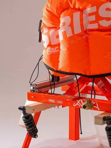 A close-up of a vibrant orange racing seat with large "SERIES" text, mounted on a sturdy frame with a shock absorber.