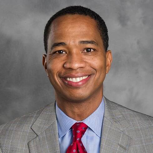 Branch Presidents: A man wearing a gray plaid suit, light blue shirt, and red patterned tie smiles at the camera, embodying the professionalism of Branch Presidents in front of a mottled gray background.