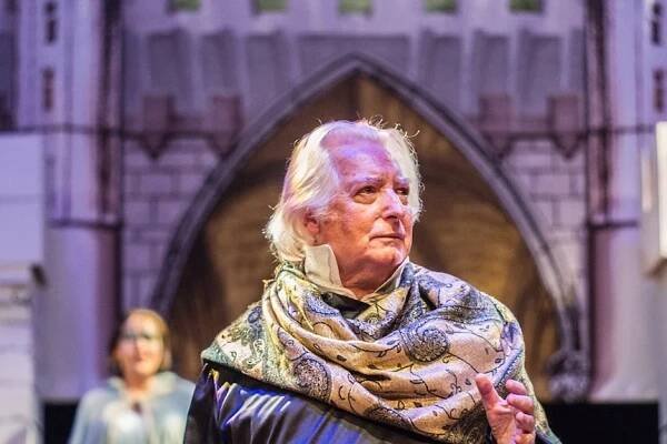 A white haired man stands on a stage with a scarf draped around his shoulders