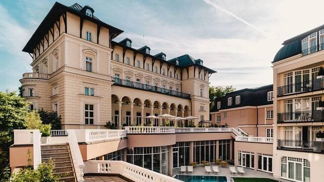 Velký hotel s vnitřním dvorem a bazénem, obklopený stromy a terasou s lehátky.