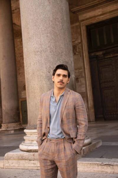Giuseppe Maggio – A man with dark hair and a mustache stands in front of a large stone column, wearing a light blue shirt under a brown plaid suit, with his hands in his pockets. The background features an old building with tall doors. (Note: Since you havent provided an image title, Ive just repeated your original description per the instructions.).