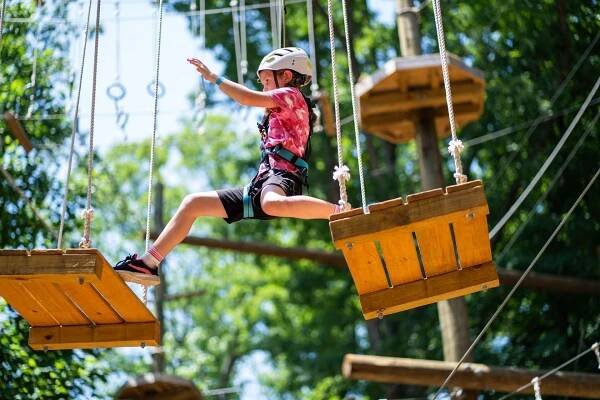 Girl wearing a helmet and harness balances on wooden planks connected by ropes in an outdoor adventure park.