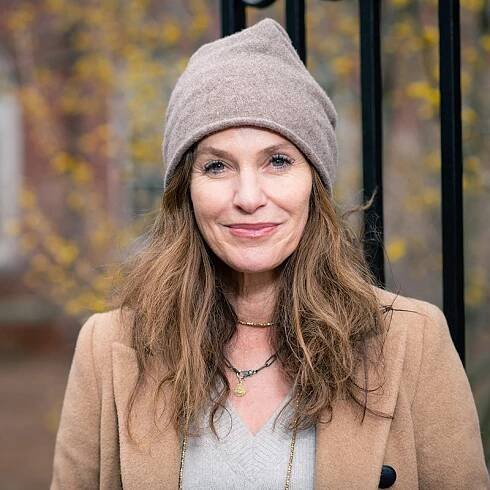 A smiling woman stands outdoors wearing a coat and a beanie.