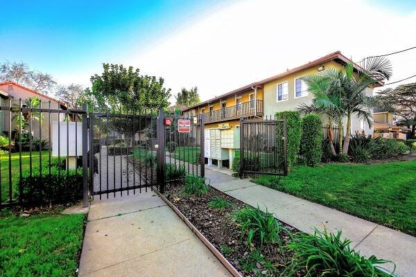 A landscaped entrance gate to an apartment community featuring a pathway lined with plants, a mailbox area on the left, and a two-story building in the background. The scene is bright and sunny, with clear blue skies and greenery surrounding the area.