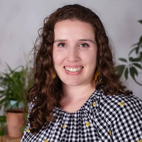 Photograph of Allison Levin, PsyD. She has a warm smile and blue eyes. Allison is wearing a black and white, gingham-checked blouse with little yellow flowers embroidered on it. Photo features a gray wall background with plants in the background, too.