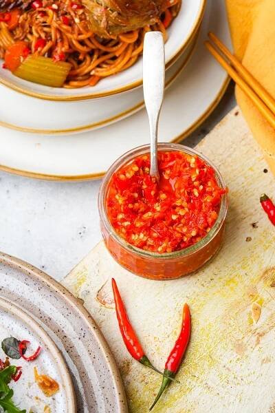 A small jar of Sambal Oelek placed on a rustic surface next to a plate of food and red chili peppers. A spoon is inside the jar. A plate of noodles is partially visible.