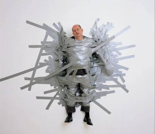 A photo of Massimo De Carlo wrapped in duct tape on a pristine white gallery wall captures the performative, absurd, and slightly cruel edge of A Perfect Day.