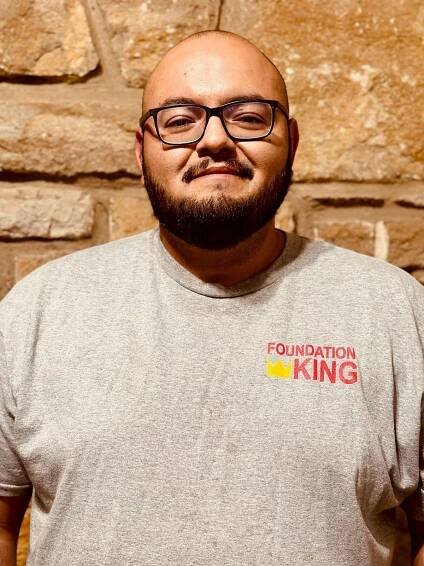 A man with a beard and glasses is wearing a foundation king t-shirt.