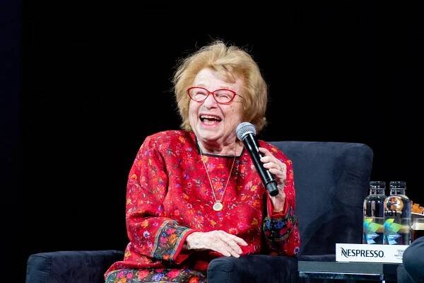 Ruth Westheimer laughing and holding a microphone. She is wearing a red floral-patterned top.