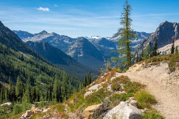 The Pacific Crest Trail winds around a high alpine ridge, with views of a deep valley and rugged North Cascades peaks in the distance.