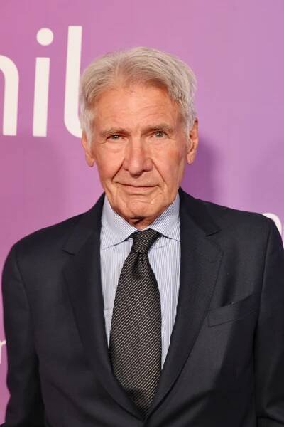 Harrison Ford in a dark suit and blue shirt with a purple background and staring at the camera.