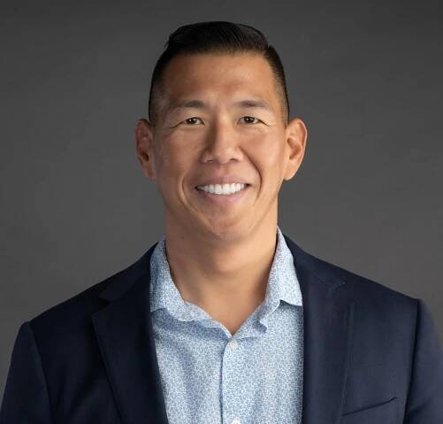 Smiling Asian man in a navy blazer and light blue patterned shirt against a gray backdrop.