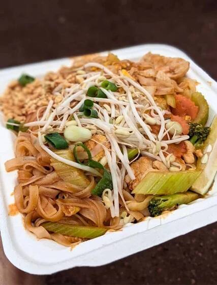 A close-up of a take-out container filled with Pad Thai, featuring noodles, bean sprouts, green onions, and various vegetables.