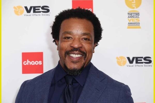 Russell Hornsby smiles in a dark blue suit.