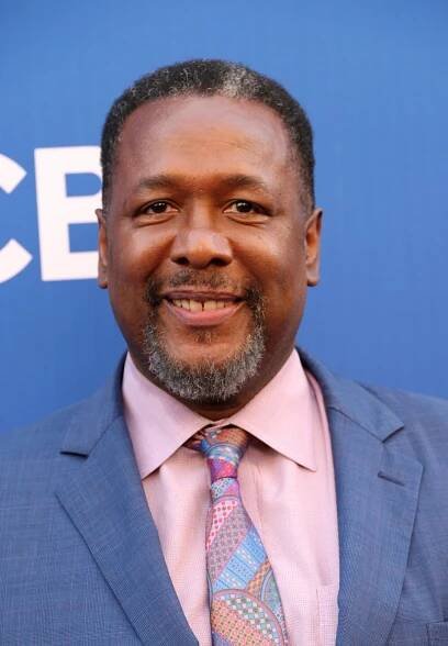 Wendell Pierce, wearing a blue jacket over a pink shirt and a colorful patterned tie, smiles on a red carpet.