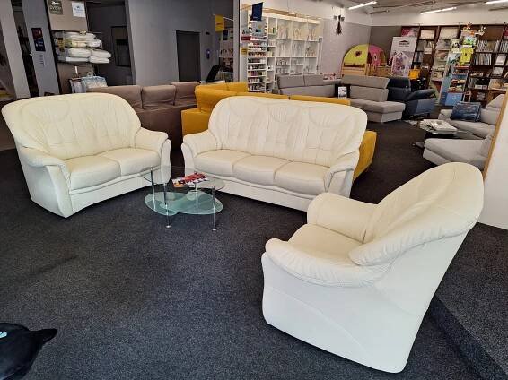 V showroomu s nábytkem je vystavena sada krémových kožených pohovek a křesla uspořádaná kolem malého skleněného konferenčního stolku na tmavém koberci, v pozadí jsou další pohovky, police a bytové dekorace.