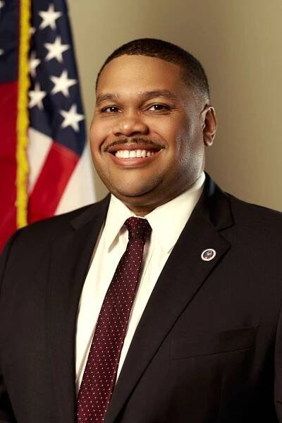 Professional headshot of government official in dark suit with burgundy tie against American flag backdrop.