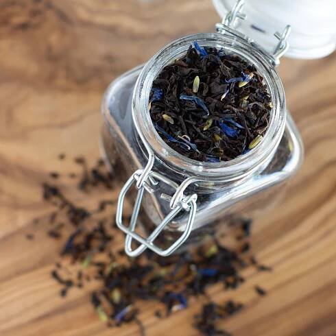 Earl Grey Lavender loose black tea leaves shown in a clear jar with tea leaves strewn around it.