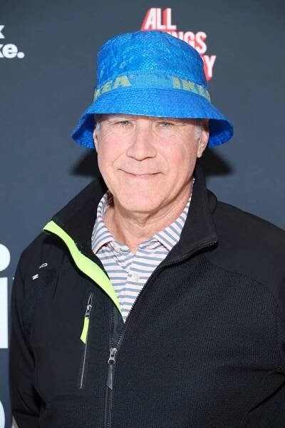 A man wearing a blue hat, striped shirt, and black jacket smiles at the camera against a press backdrop