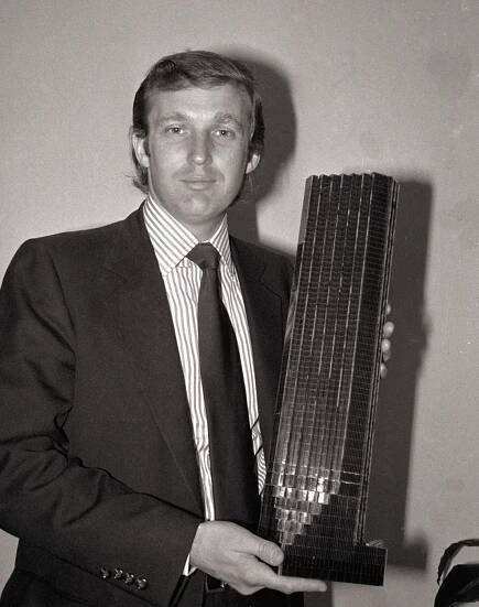 A black and white photo of a young Donald Trump wearing a suit and holding a model of a tall building.